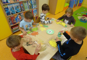 Sześcioro dzieci siedzi przy stole na którym rozłożone są kolorowe liście papierowe oraz owoce. Dzieci dobierają owoce do liści drzew.